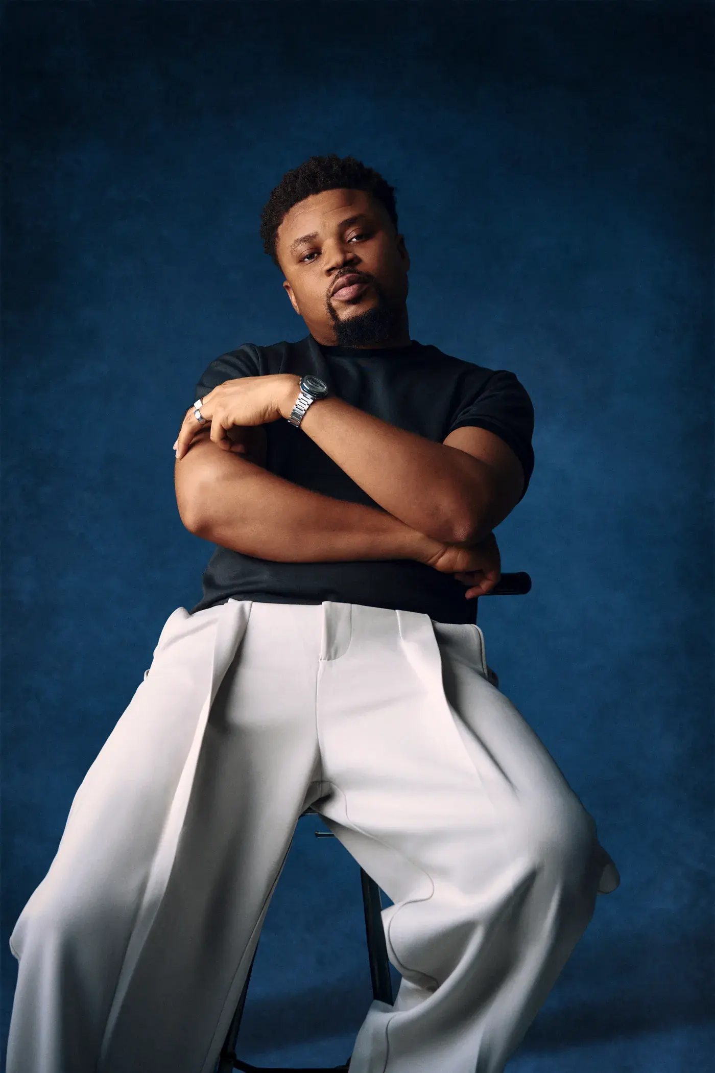 Ko'rale seated with his arms crossed, black tee and grey trousers, deep blue backdrop