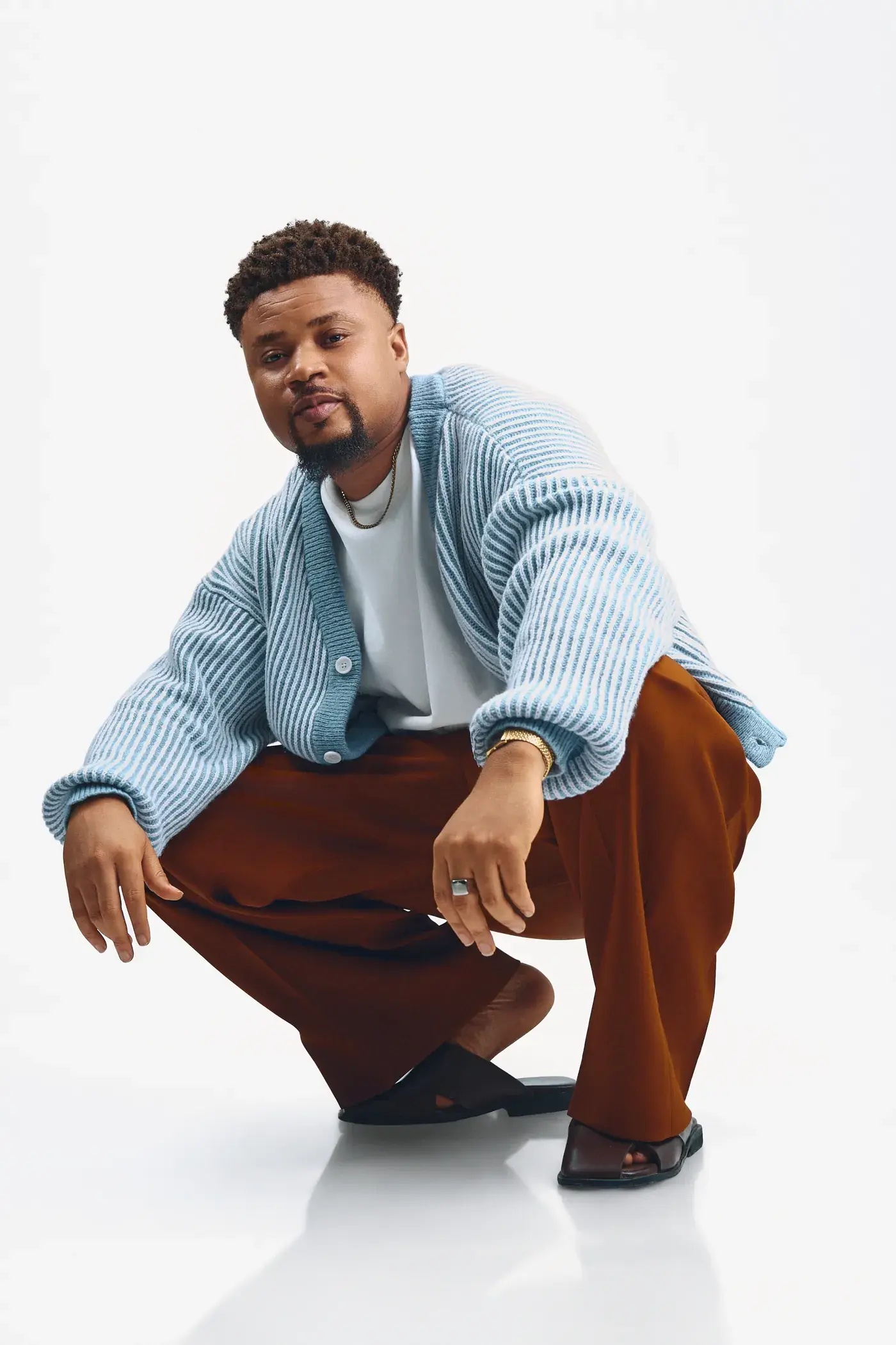 Ko'rale crouching and looking into the camera in a blue cardigan and brown trousers on white