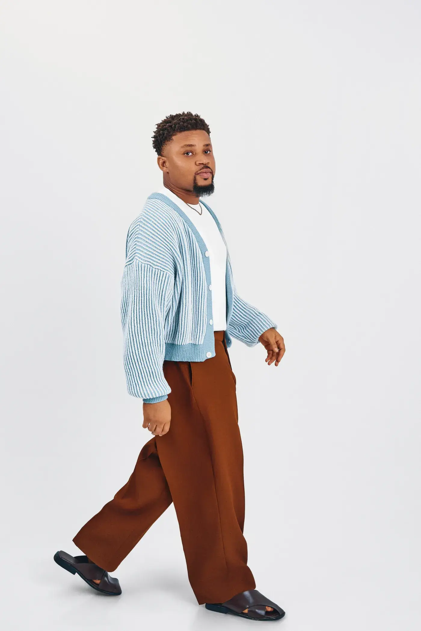 Ko'rale walking in profile in a blue cardigan and brown trousers on white